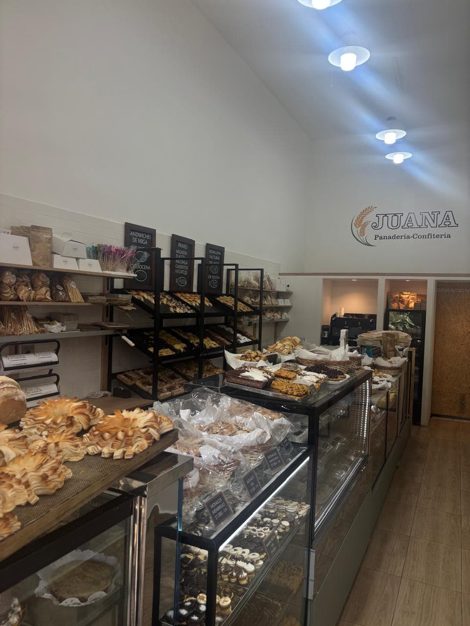 Interior de Panadería Juana: estantes con panes y facturas, pizarras de precios, mostrador vidriado y el logo de Juana en la pared